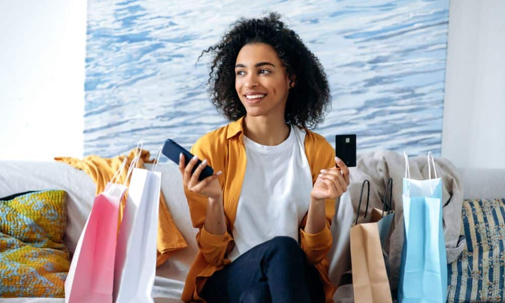 A photo of a woman holding her phone and credit card with shopping bags beside her.