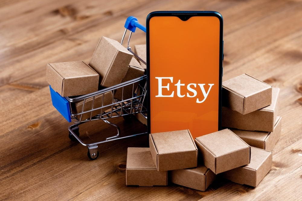 Etsy app on phone surrounded by mini shopping cart and carton boxes.