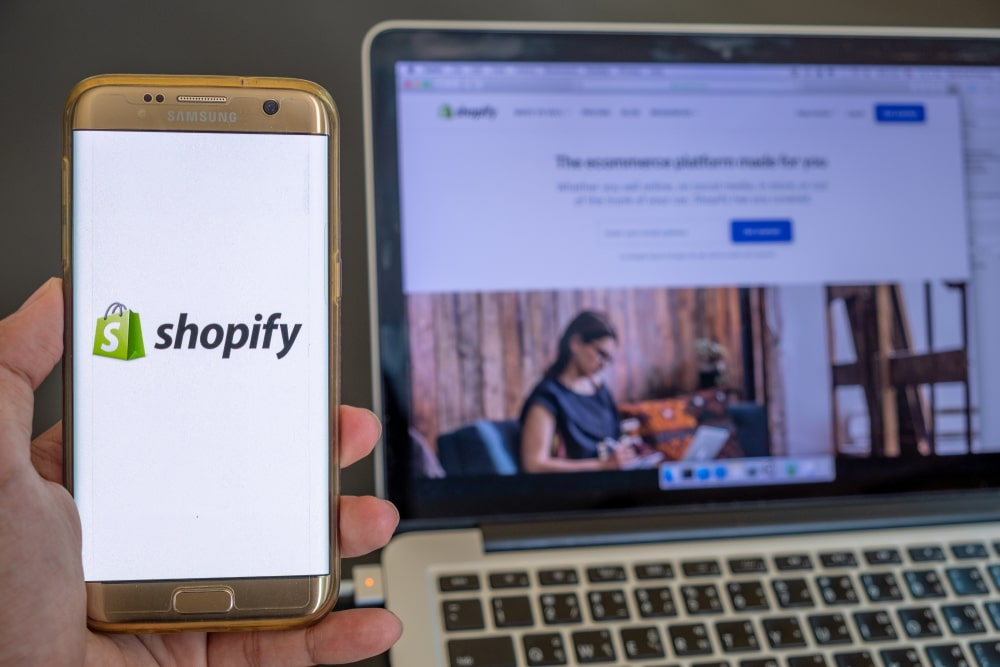 Phone running the Shopify app against a blurred laptop background.