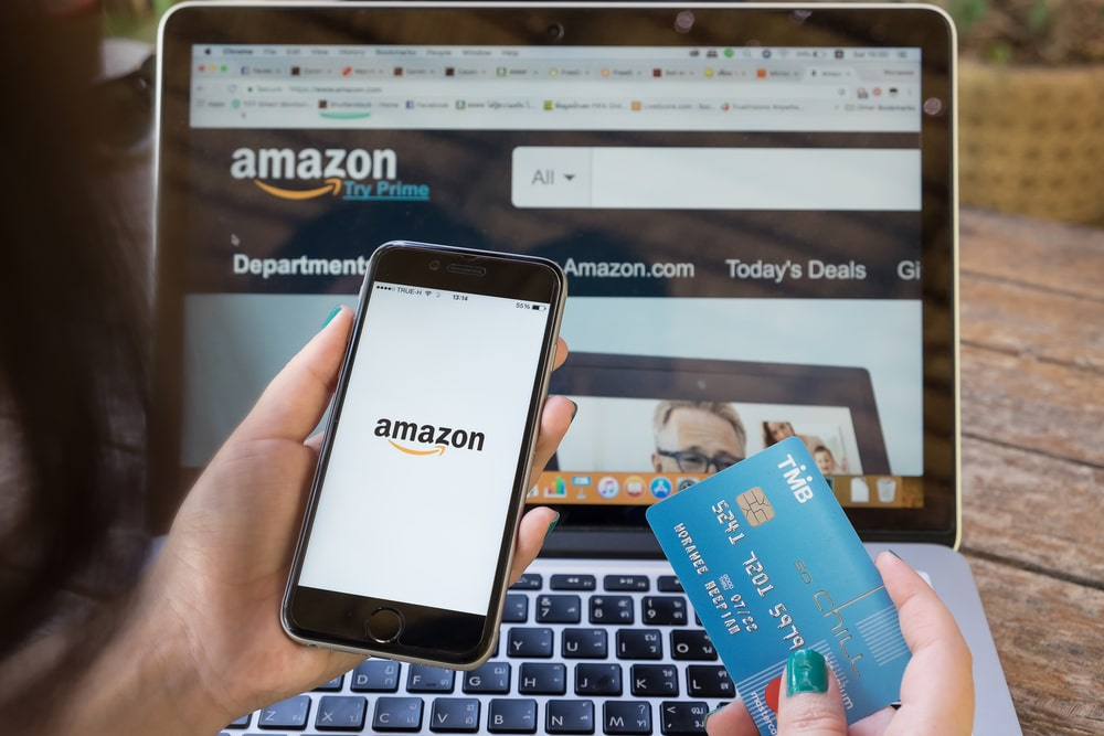 une main tenant un téléphone avec le logo Amazon, une carte de crédit et un ordinateur portable avec le site web d'Amazon à l'écran