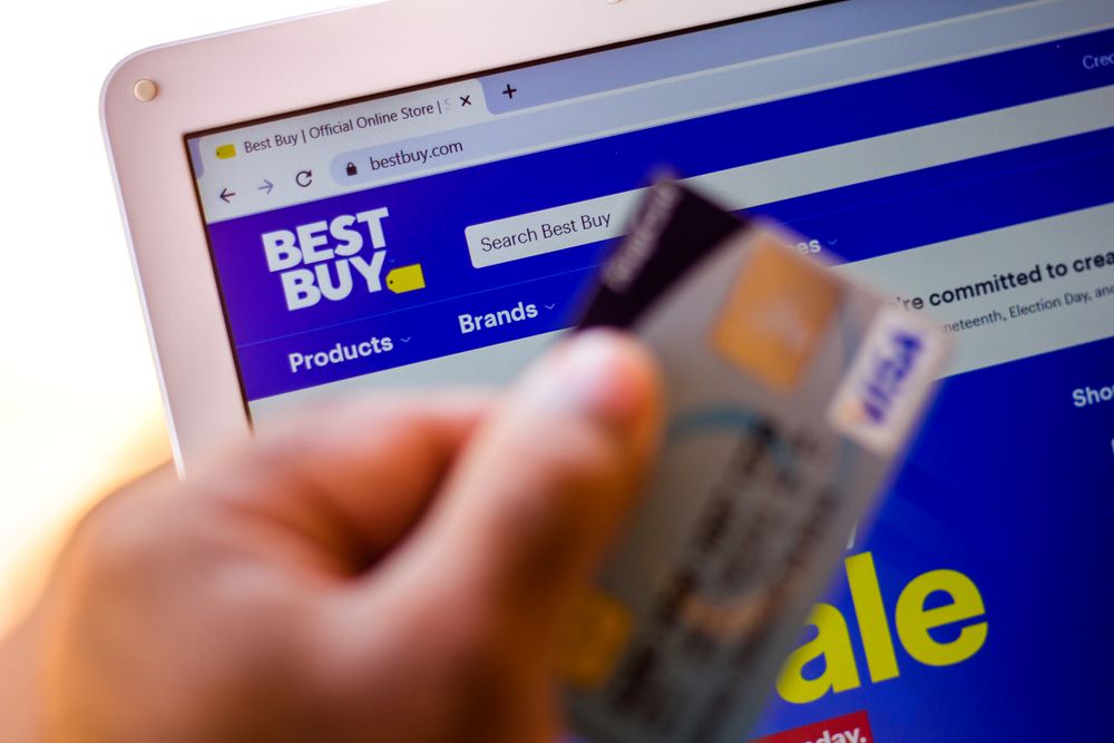 A hand holding up a card in front of a laptop with Best Buy site on screen