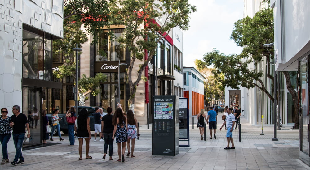 A sunlit outdoor shopping avenue lined with modern storefronts including a Cartier store, shoppers walking, and a directory map on the sidewalk.