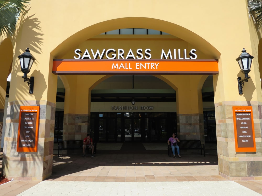 Entrance of Sawgrass Mills Mall with an orange sign above the doorway and two directory boards on either side listing various fashion stores. Two people are sitting on a bench near the entrance.
