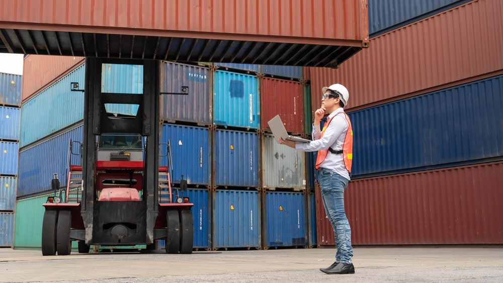 A dockworker observing a forklift carrying a shipping container.