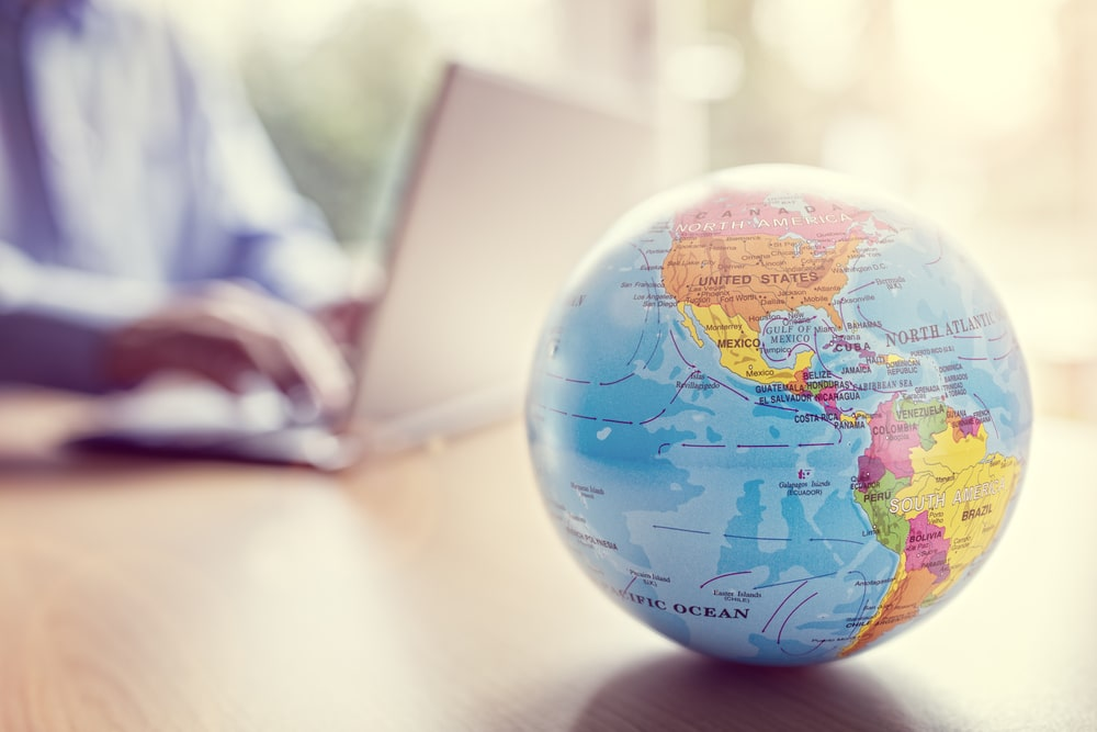 A person using a laptop with a small globe on the desk in front of them, with the focus on the globe showing North and South America.