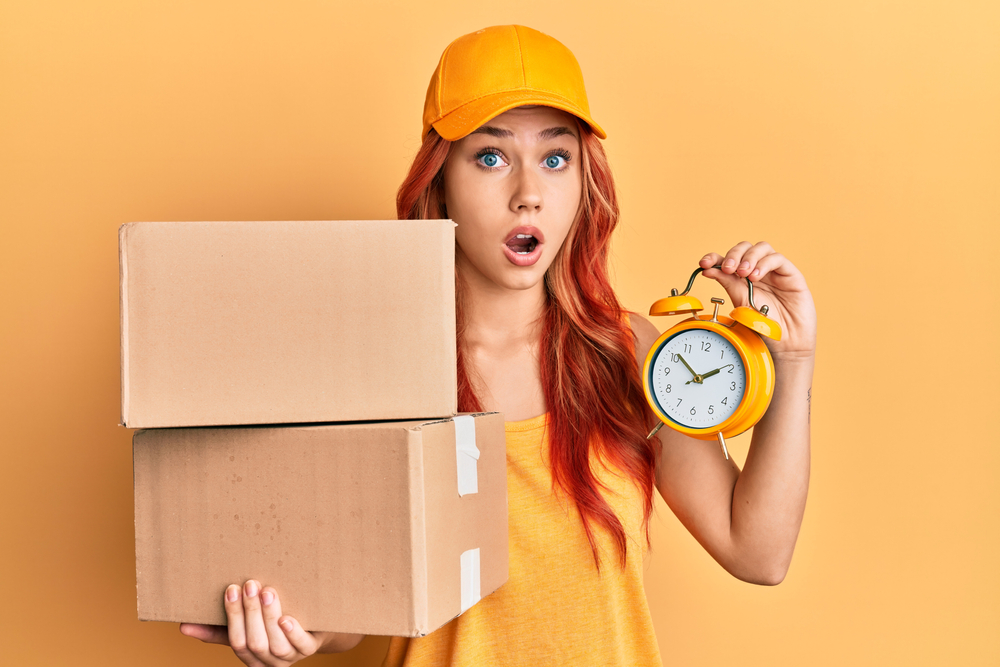 A woman wearing an orange tank top and orang cap carrying an orang alarm clock on the right hand and two boxes on the left hand