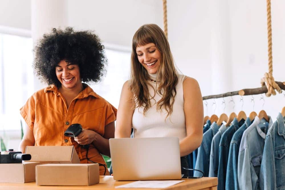 Smiling business women preparing packages while using a laptop, with hanging clothes at their back.