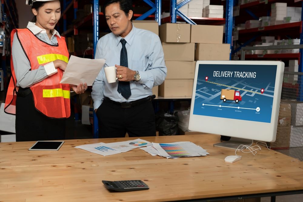 A man and woman looking at a document and a monitor with delivery tracking on the screen