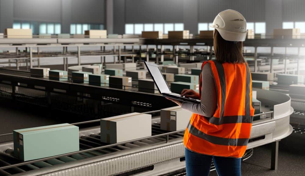 A woman with a laptop facing packages on a warehouse carousel